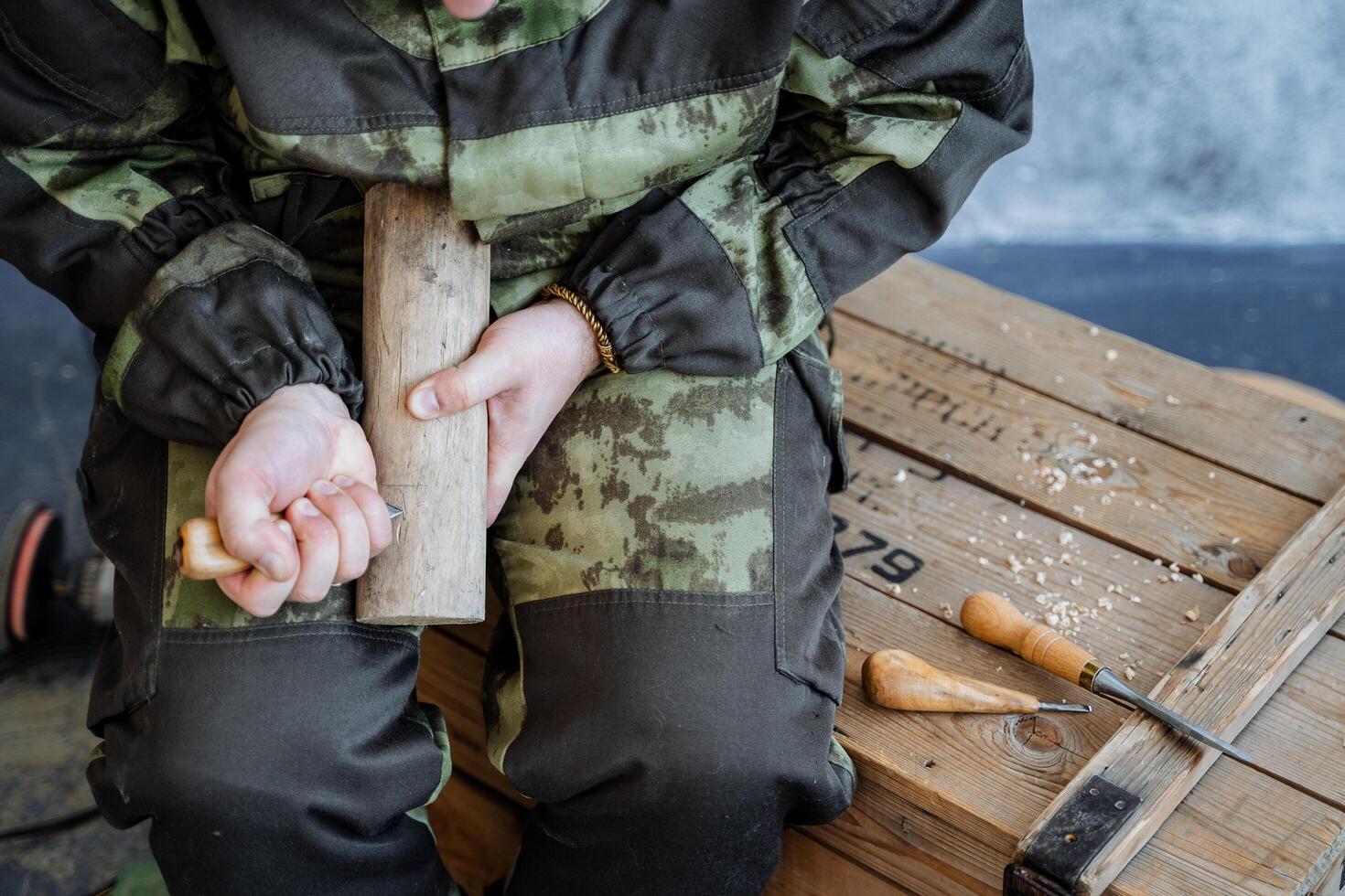 A man in camouflage uniform carves parts in wood. Wood processing and creation of toys and souvenirs. Around on a wooden box are tools. photo