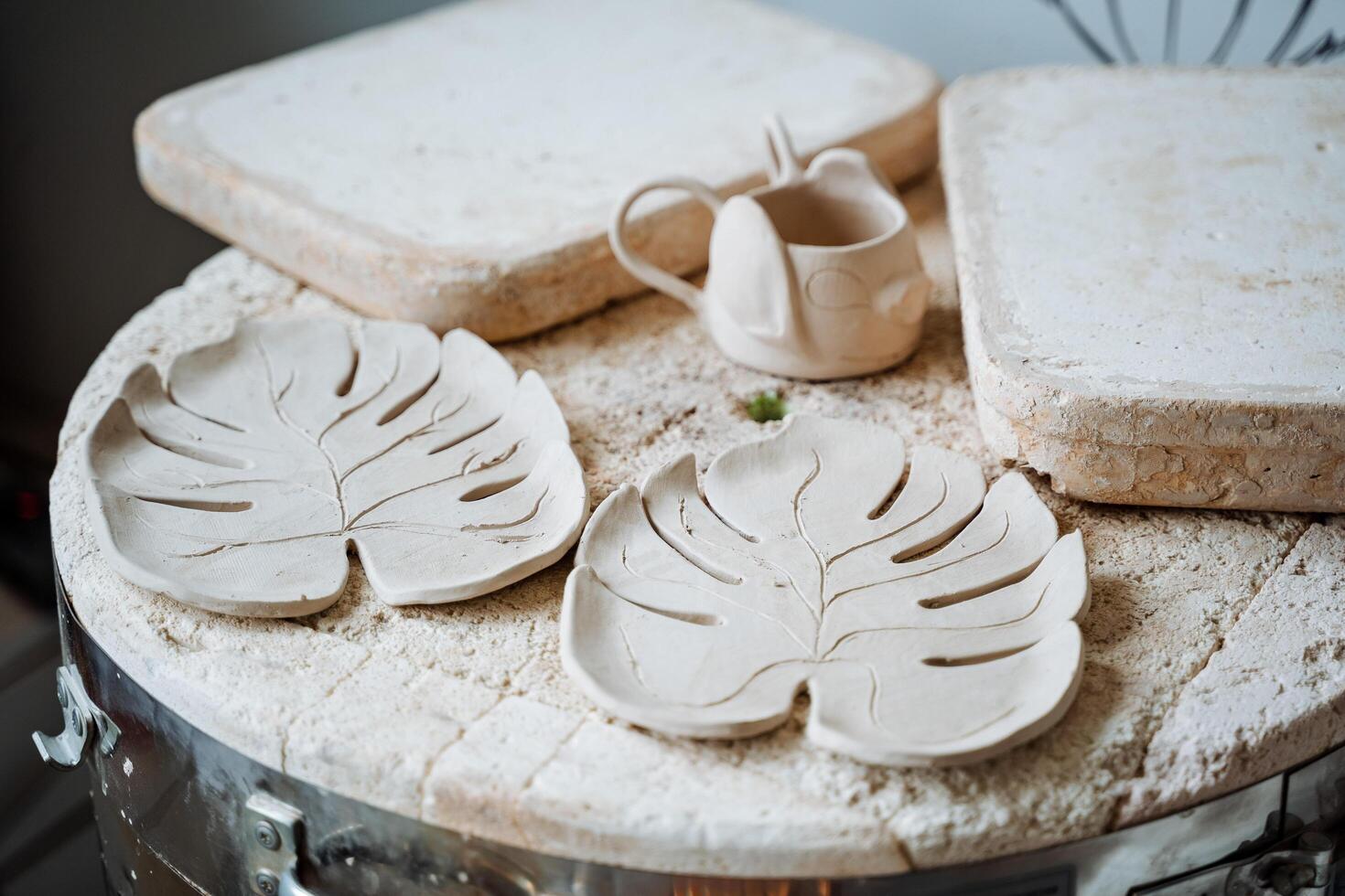 Plates in the form of monstera leaves made of clay on a wooden table in a pottery studio. Nearby are two boards and a decorative mug.Creative activities and the creation of ceramics. photo
