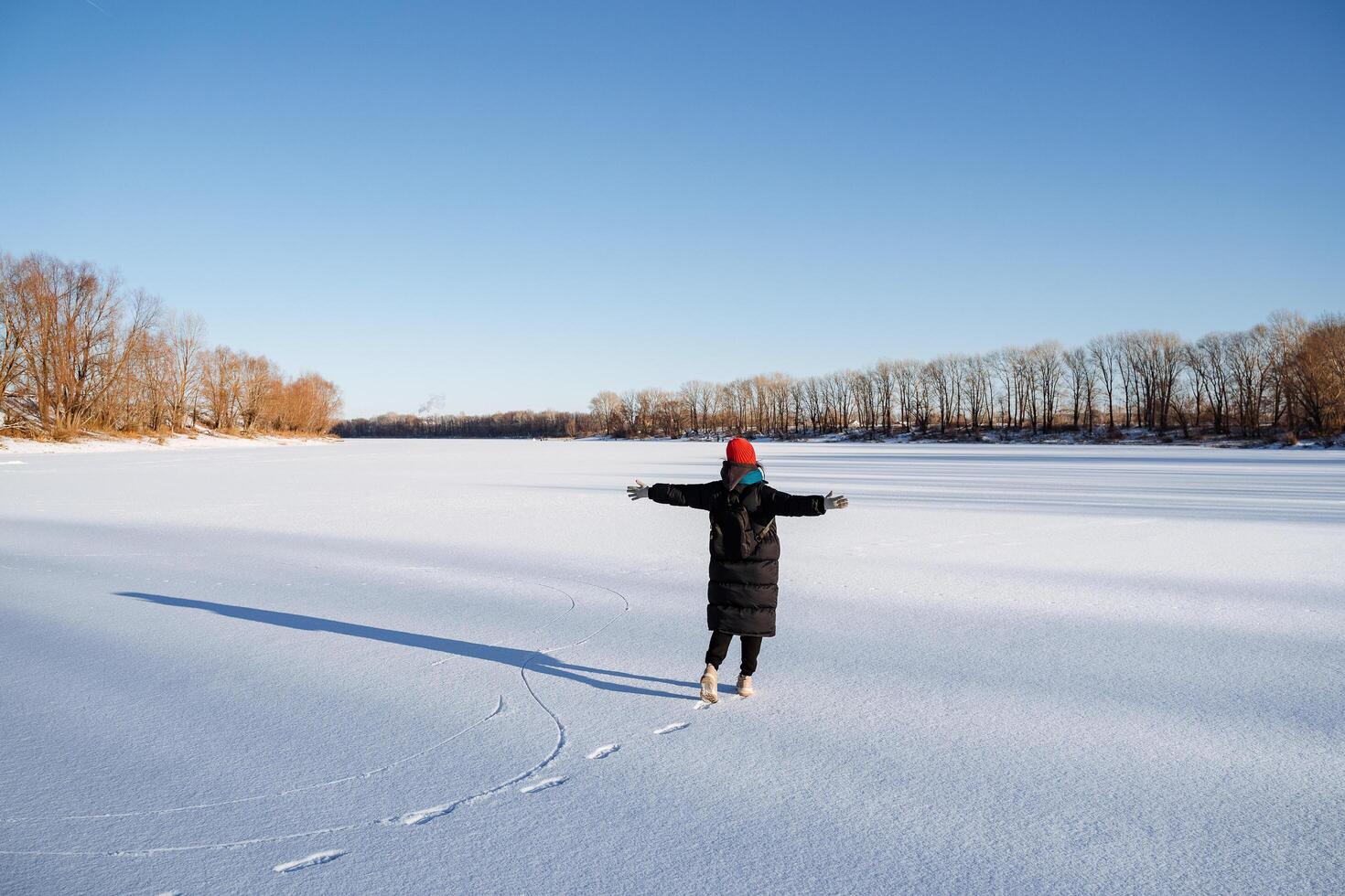 A young girl in a bright hat walks on a frozen lake. Delight and joy from the winter landscape with bright sun, clear sky. Walk in nature. photo