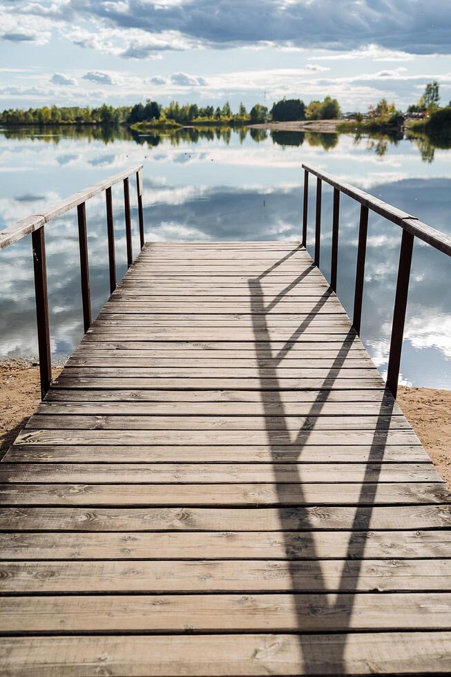 A close shoot of a wooden pier by the blue clear water, surrounded by forest on a sunny day. Solar glare on the water. Outdoor recreation by the lake. Clouds reflect in the water photo