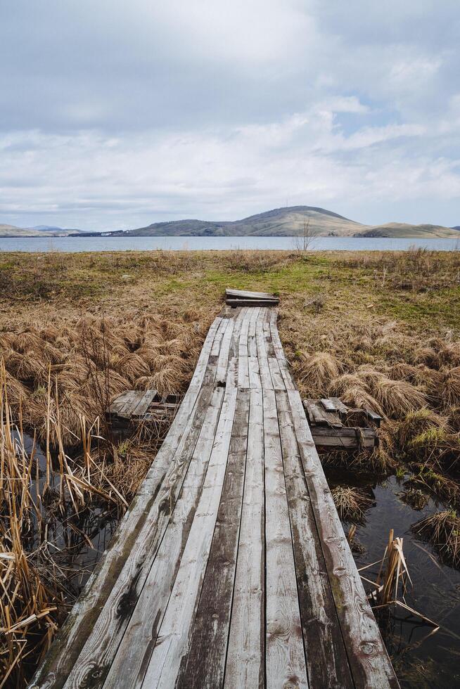 un antiguo de madera puente yendo Derecho a el agua mediante pantanoso suelo con seco césped. oscuro y obeso nubes en el cielo, adelante de el lago subir pequeño montañas. foto