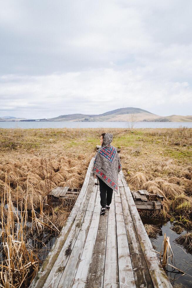 un joven mujer en un de colores poncho camina a lo largo un antiguo de madera puente mediante un campo con un pantano. el puente va Derecho a el lago, detrás cuales el montañas elevar. paisaje en lago bannoye foto