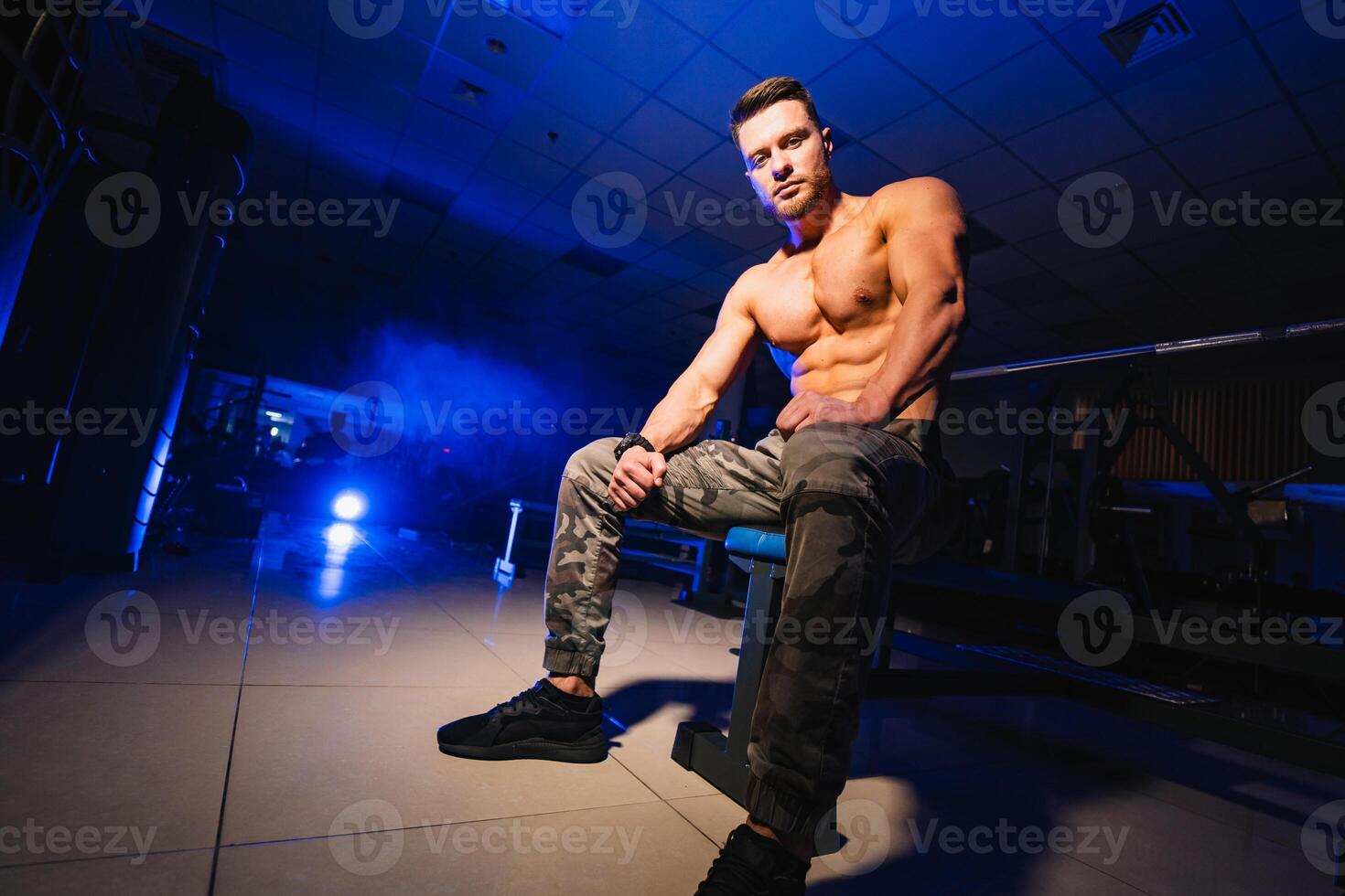 Man rests in gym after having a workout. Strong male with perfect body in the dark background with smoke. Closeup. photo