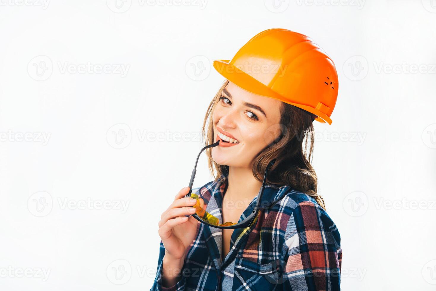 Portrait of passionate female builder with helmet and glasses touching her lips looking at camera and smiling. Sexy woman in protective helmet, glasses holding in hand. photo