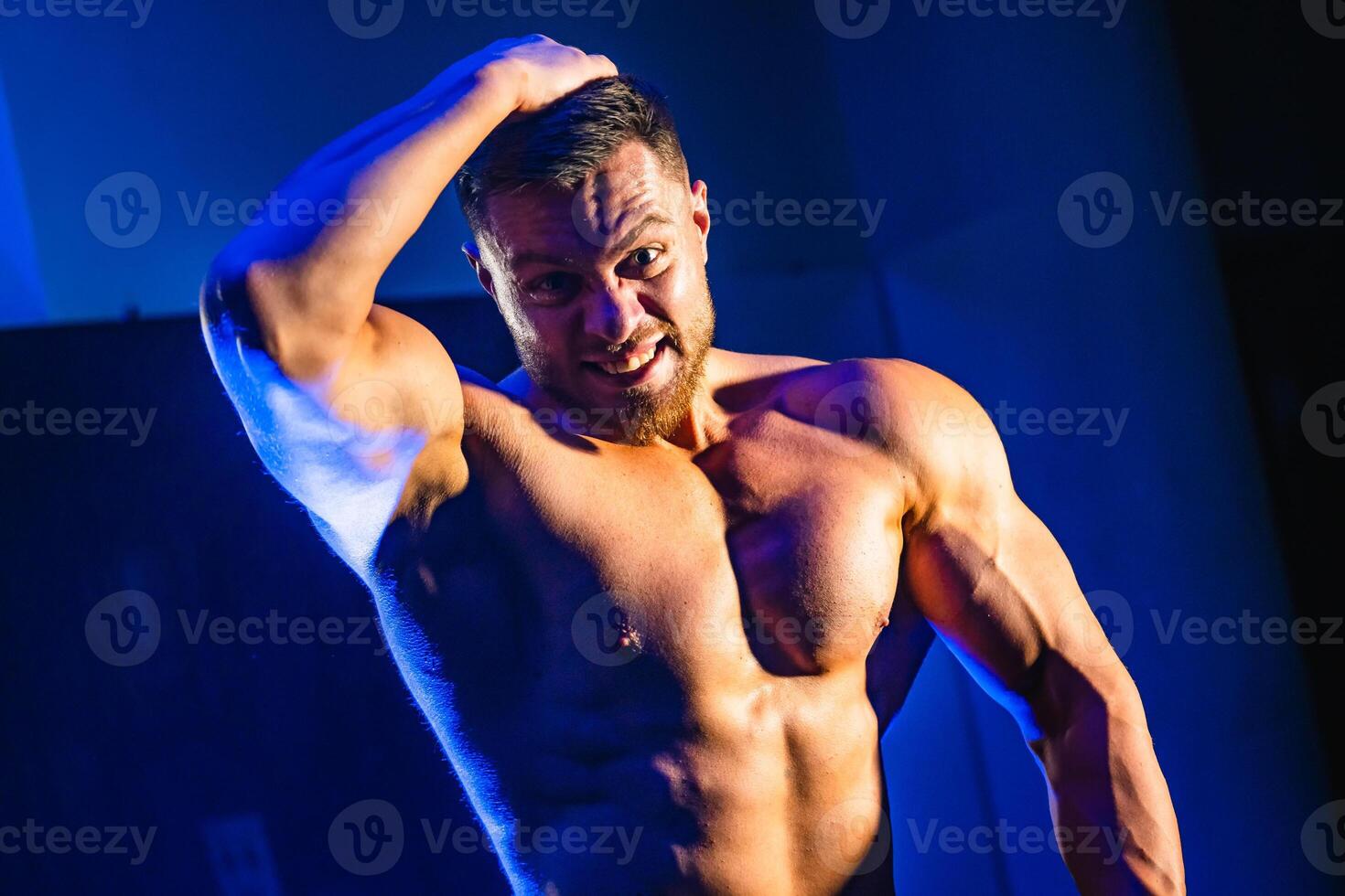 hermoso hombre con grande músculos, posando a el cámara en el gimnasia, negro y azul antecedentes. retrato de un sonriente carrocero. de cerca. foto