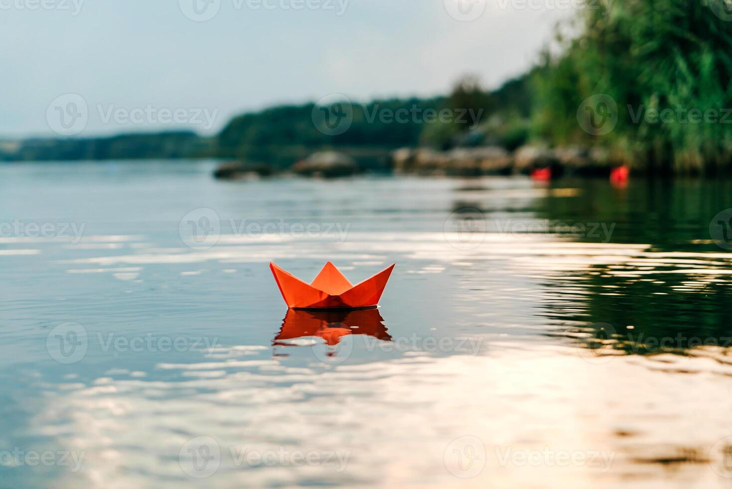 Red paper origami boat floats on the surface of the water and reflects itself on the natural background. Paper ship sailing on blue water surface. photo