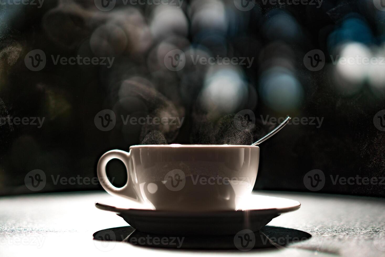 White cup with hot liquid and steam on black blurred background. Teaspoon in a cup. Tea and coffee concept. photo