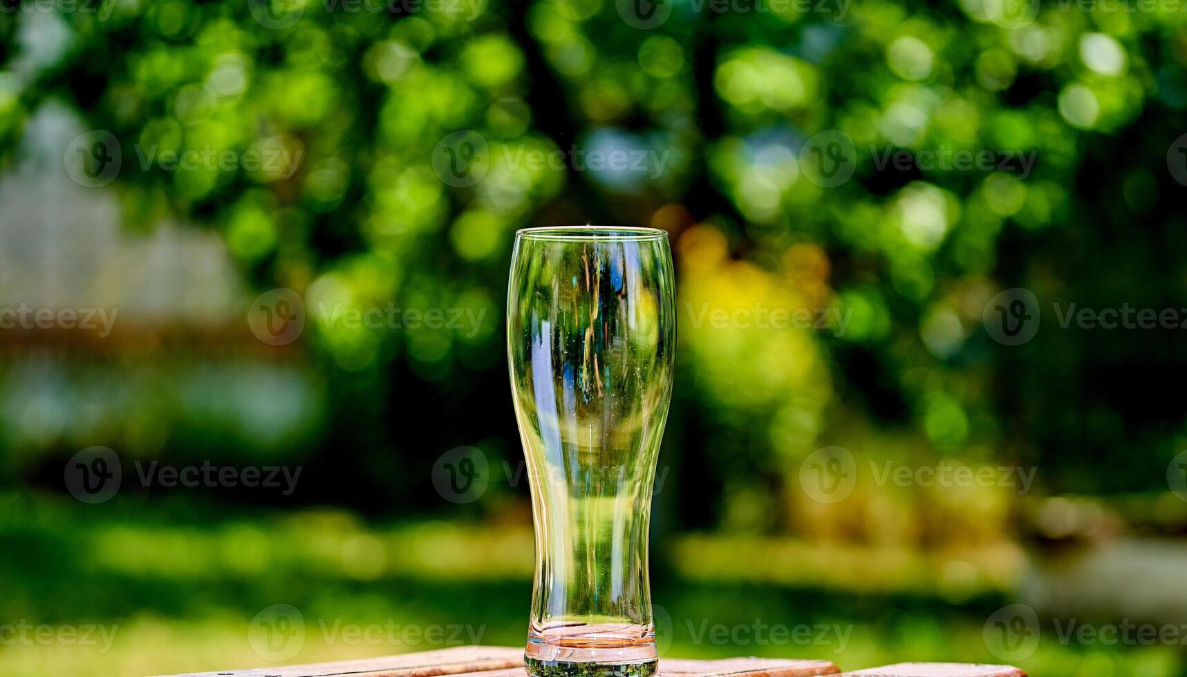 Close-up picture of an ampty beer glass on a brown wooden table. Sun shines through the glass. Blurred green nature background. photo