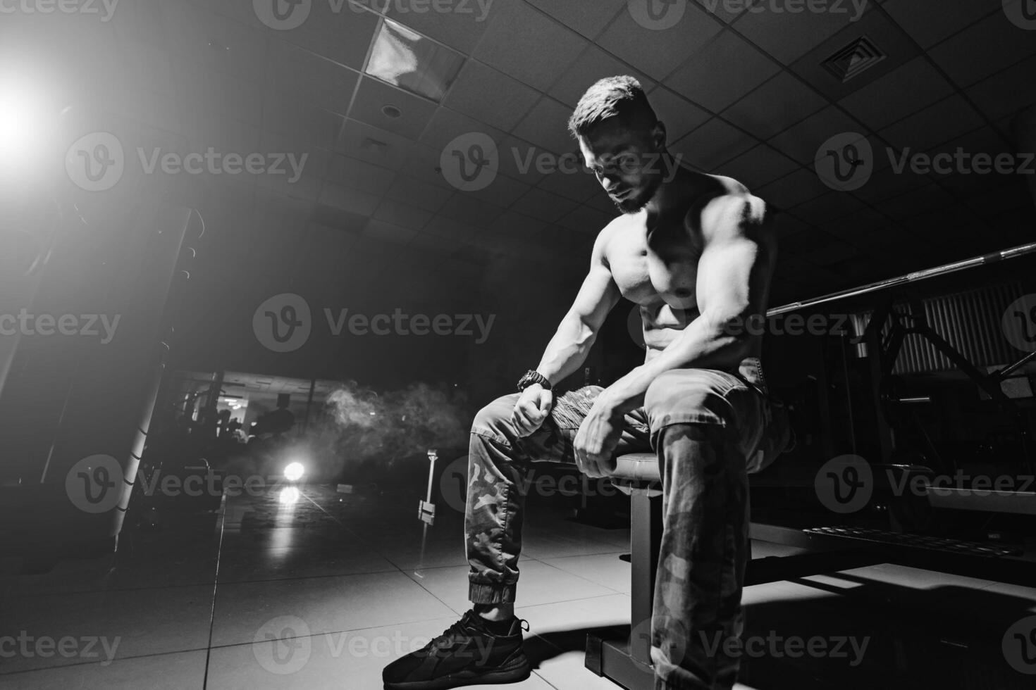 Man rests in gym after having a workout. Storng male with perfect body in the dark background with smoke. Closeup. photo