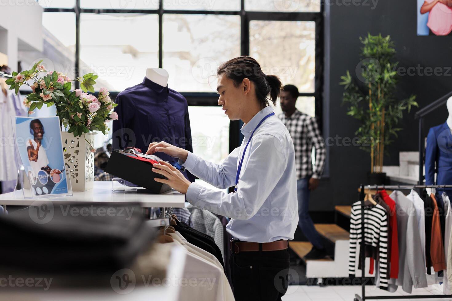 Asian manager arranging elegant tie and clothes, preparing clothing store for opening. Employee working with fashionable merchandise and accessories, checking new fashion collection in modern boutique photo