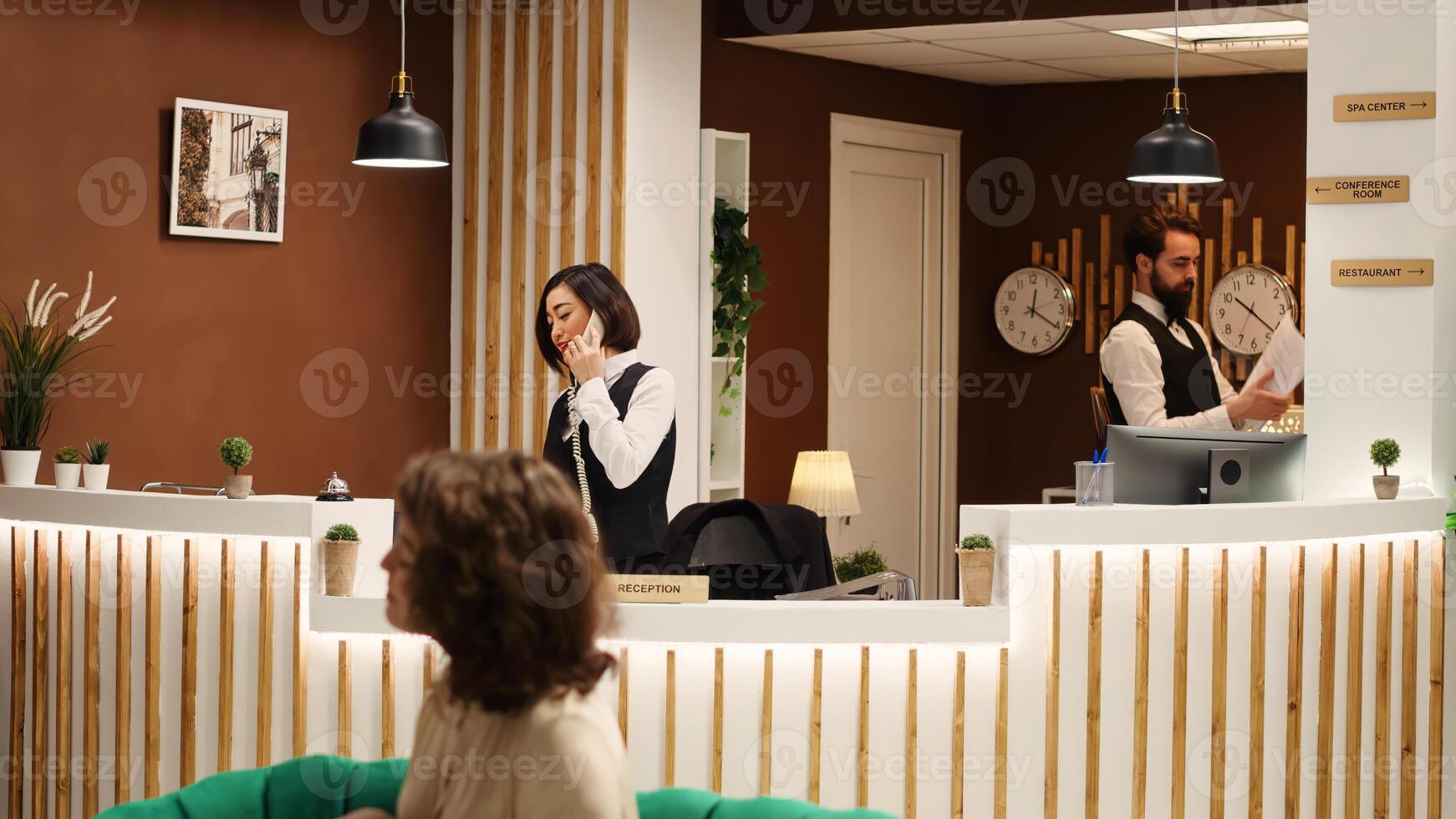 Smiling asian receptionist and coworker verifying resort reservations info while working in stylish hotel lobby. Concierge personnel assisting incoming guests with accommodation details photo