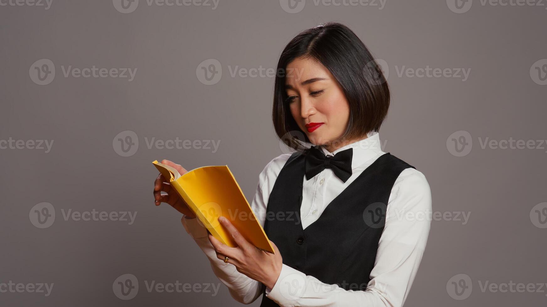 Asian receptionist reading a literature book over grey background, enjoying lecture hobby with a novel story in studio. Hotel concierge employee reads a fiction tale for knowledge. Camera B. photo