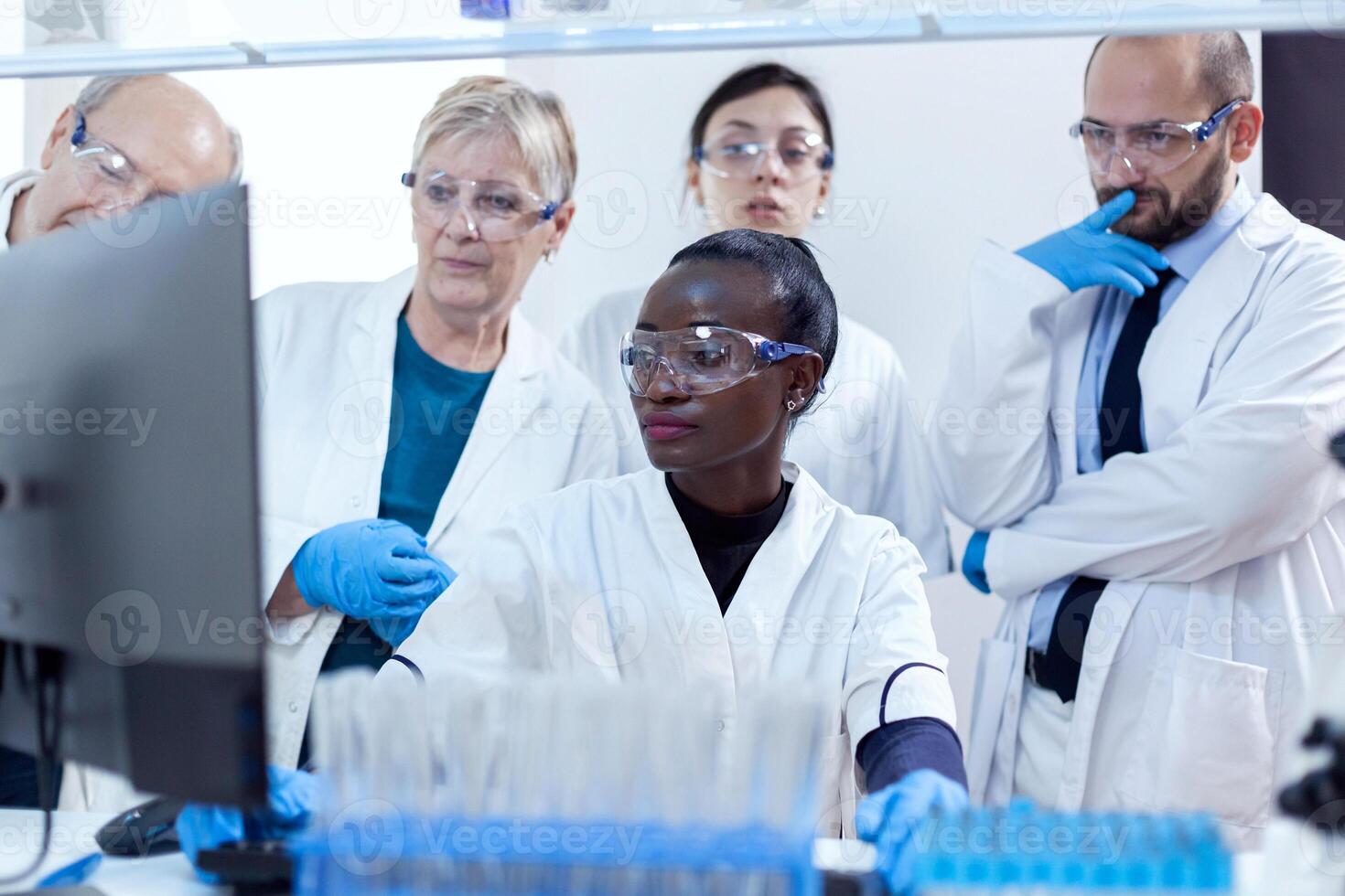 Multiethnic group of laboratory scientists discussing their research in front of computer. African healthcare researcher in biochemistry laboratory wearing sterile equipment. photo