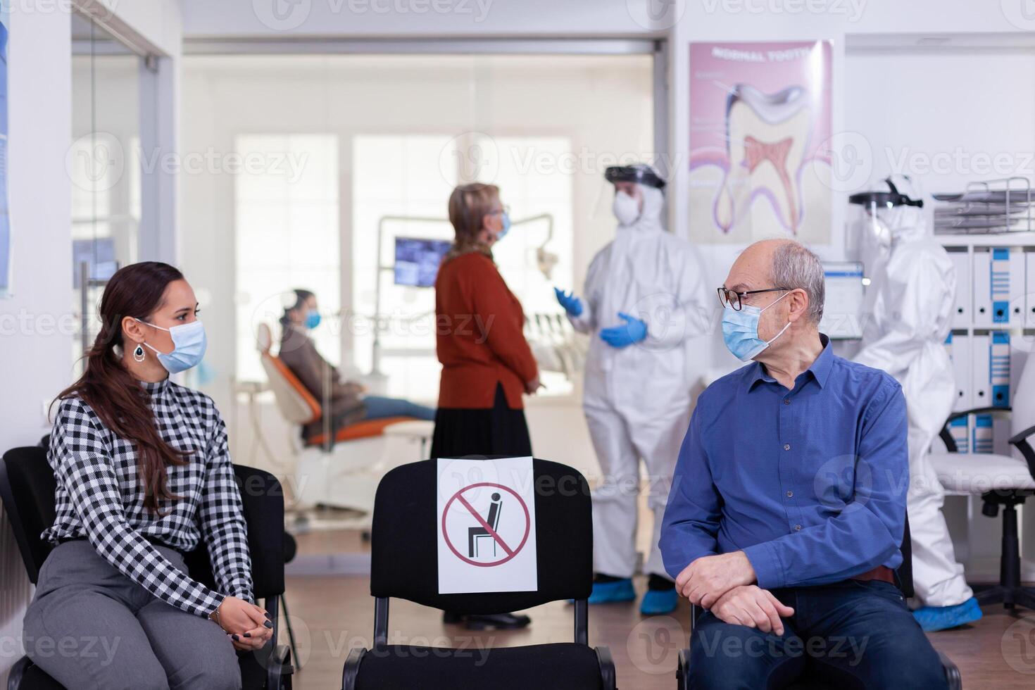 Senior man with face mask discussing with woman patient in stomatology clinic in waiting room, keeping social distancing during global pandemic with coronavirus. Doctor standing wearing ppe suit. photo