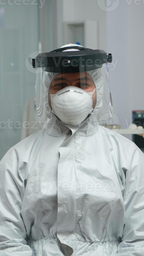 Dentist in coverall looking and smiling at camera sitting in stomatological clinic during pandemic coronavirus. Orthodontic on video call wearing protection suit, face shield, mask and gloves. photo