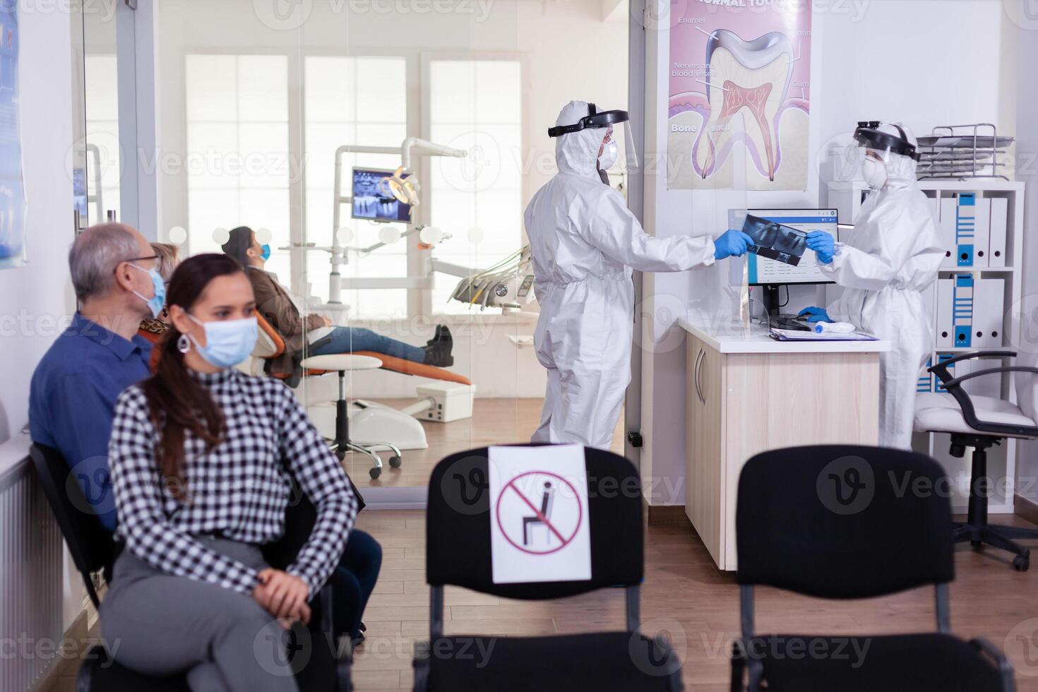 Stomatology dentist dressed in ppe suit taking patient x-ray from receptionist in clinic hallway, keeping social distancing during global pandemic with coronavirus. photo