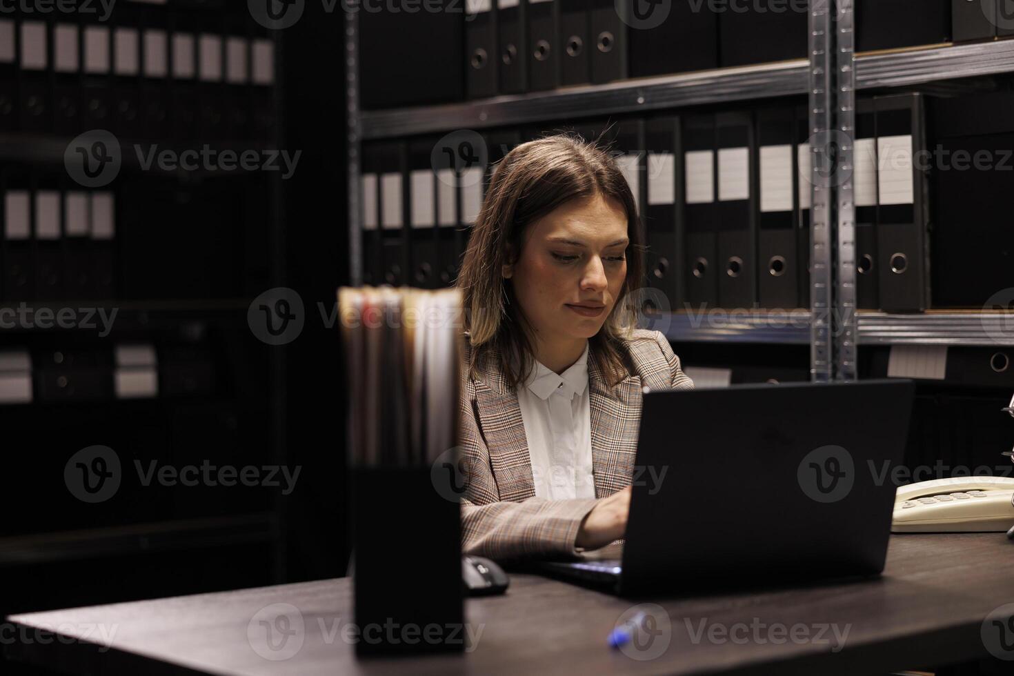 Bookkeeper analyzing administrative files on laptop computer, searching for bureaucracy record in corporate depository. Manager in formal suit working at accountancy report in storage room photo