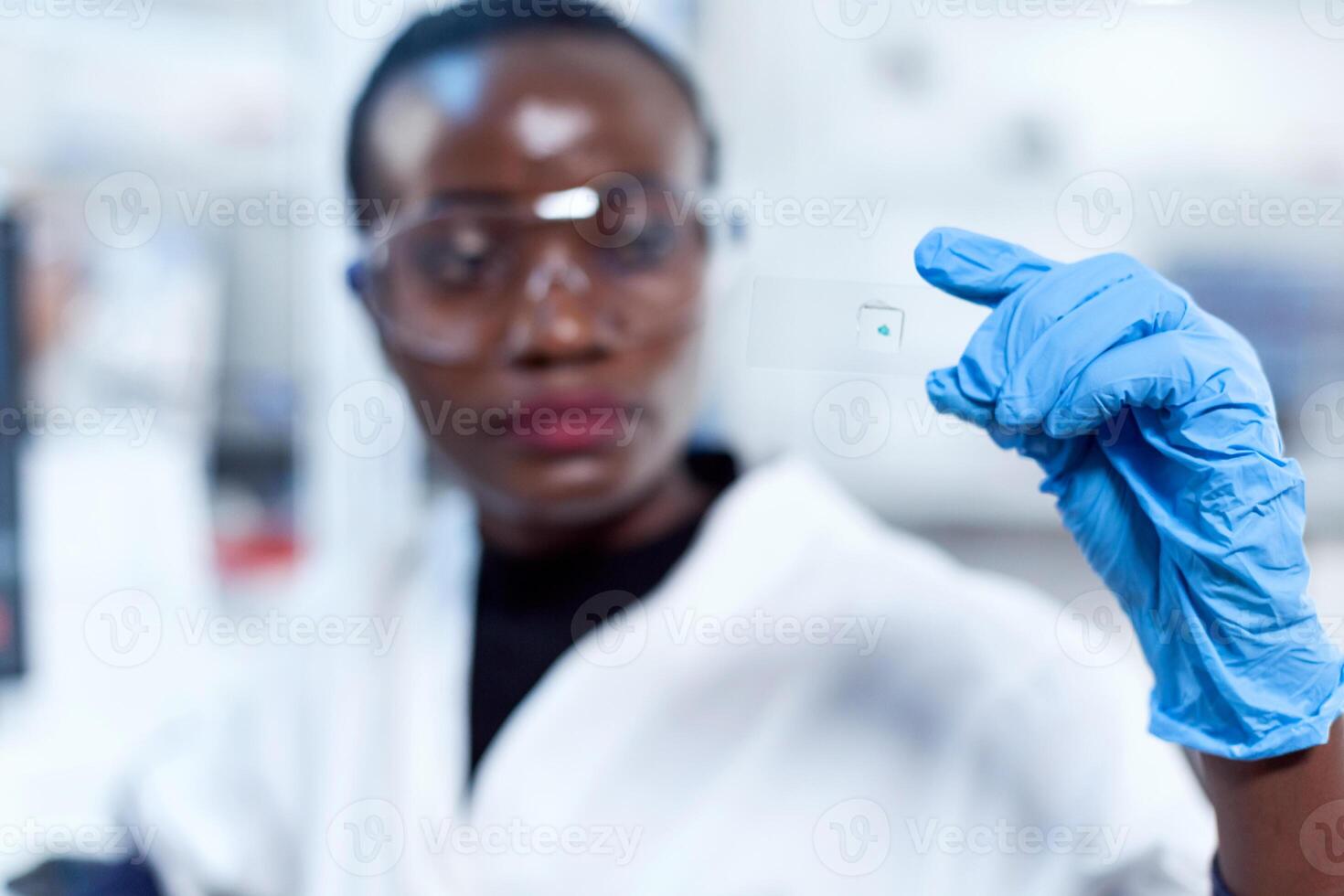 Sample glass slide in focus while chemist is looking at it during lab investigation. African healthcare scientist in biochemistry laboratory wearing sterile equipment. photo