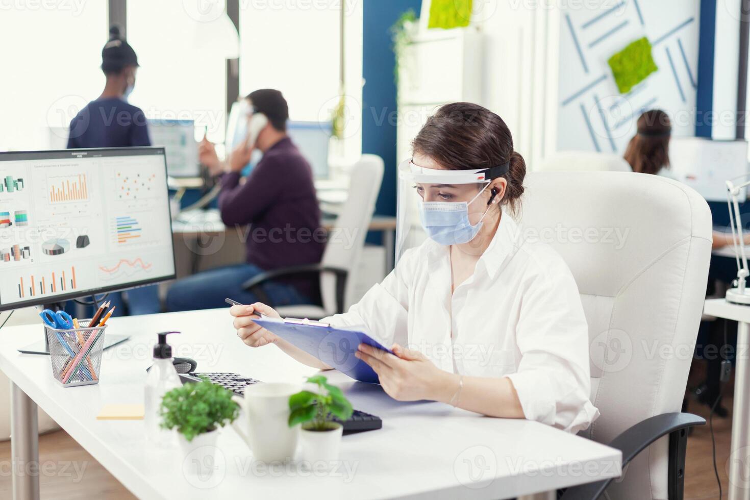 Entrepreneur wearing face mask against covid19 using wireless headphones for remote call. Employees with visors working in corporate company workspace respecting social distance analysing data and graphs. photo