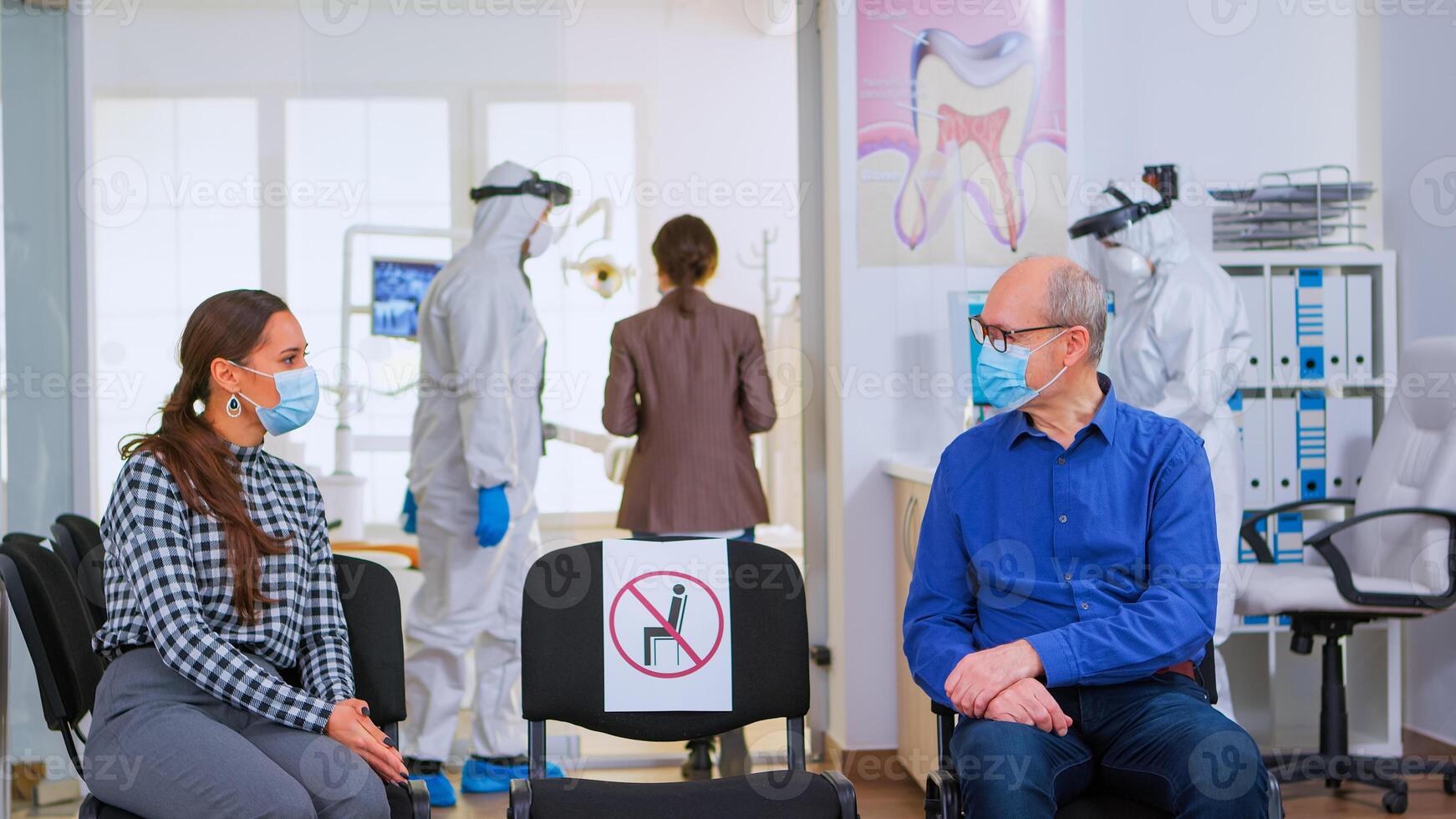 People with protection masks discussing sitting on chairs keeping social distance in stomatological clinic, waiting for dental doctor during covid-19 pandemic. Concept of new normal dentist visit photo