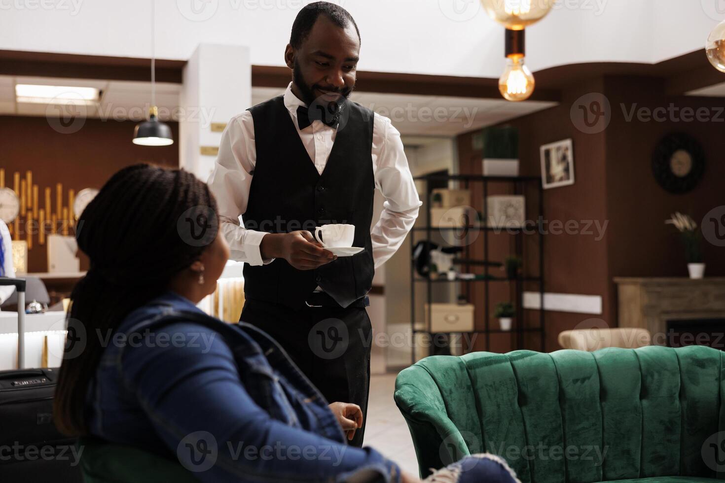 Pleasant smiling hotel worker offering free coffee to tired guest waiting for check-in process in lobby, waiter serving cappuccino to tourist arriving at resort. Customer service in hospitality photo