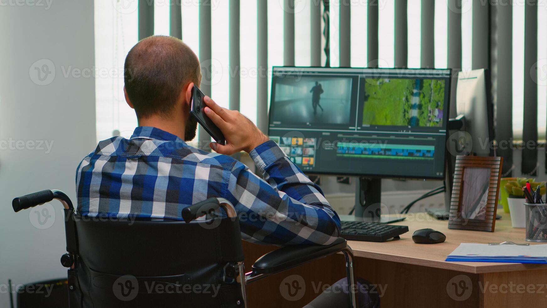 Disabled freelancer videographer talking on phone during editing a video project creating content sitting in wheelchair in modern company office. Creator blogger working from photo studio.