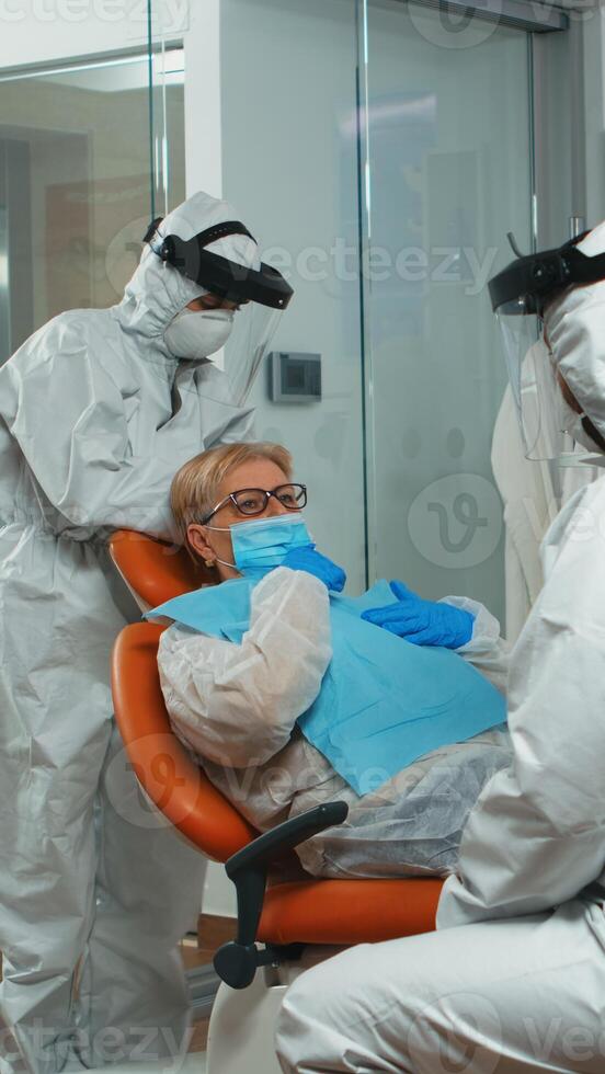 Dentist nurse in coverall putting dental bib to old woman before stomatological examination during covid-19 pandemic. Concept of new normal dentist visit in coronavirus outbreak wearing protective suit and face shield photo