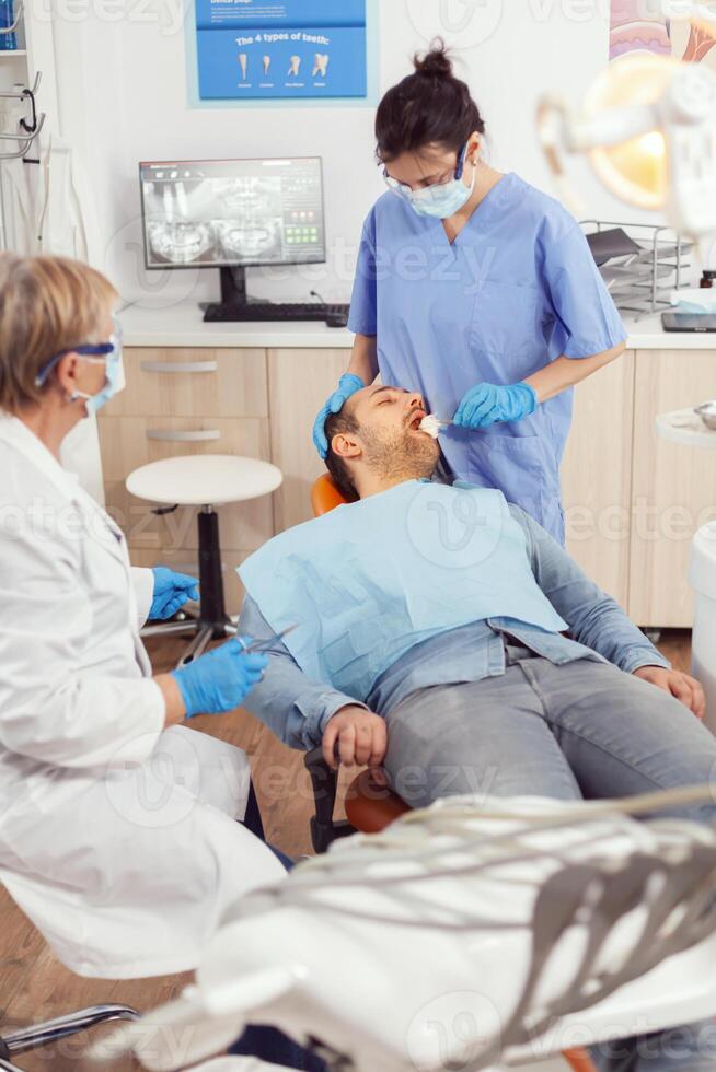 Stomatologist nurse looking in patient mouth preparing for dental surgery. Dentist asisstant with face mask examining sick man with dental bib before intervention sitting on stomatological chair photo