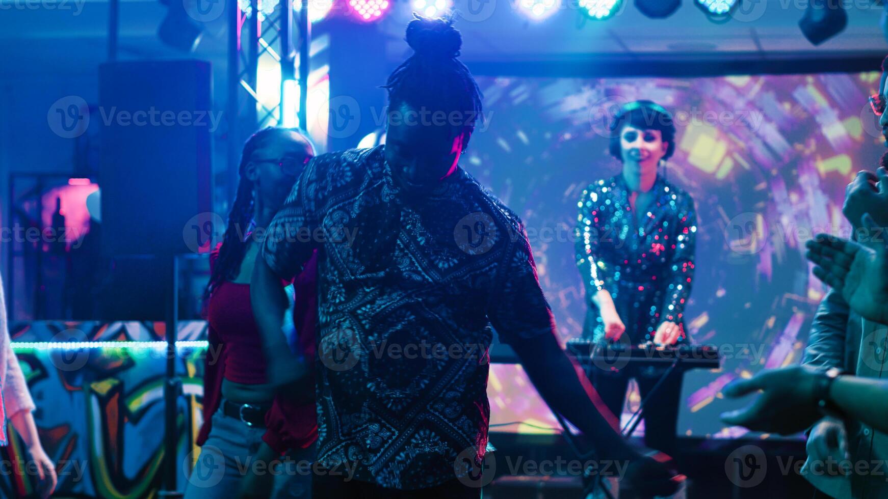 Group of friends dancing in the club, enjoying live DJ performance on colorful dance floor. Young funky people having fun at nightclub with disco lights and alcohol, clubbing. Tripod shot. photo