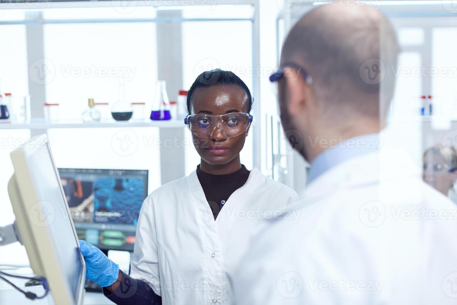 African scientist discussing virus trial with virolog in chemistry laboratory. Multiethnic team of medical researchers working together in sterile lab wearing protection glasses and gloves. photo
