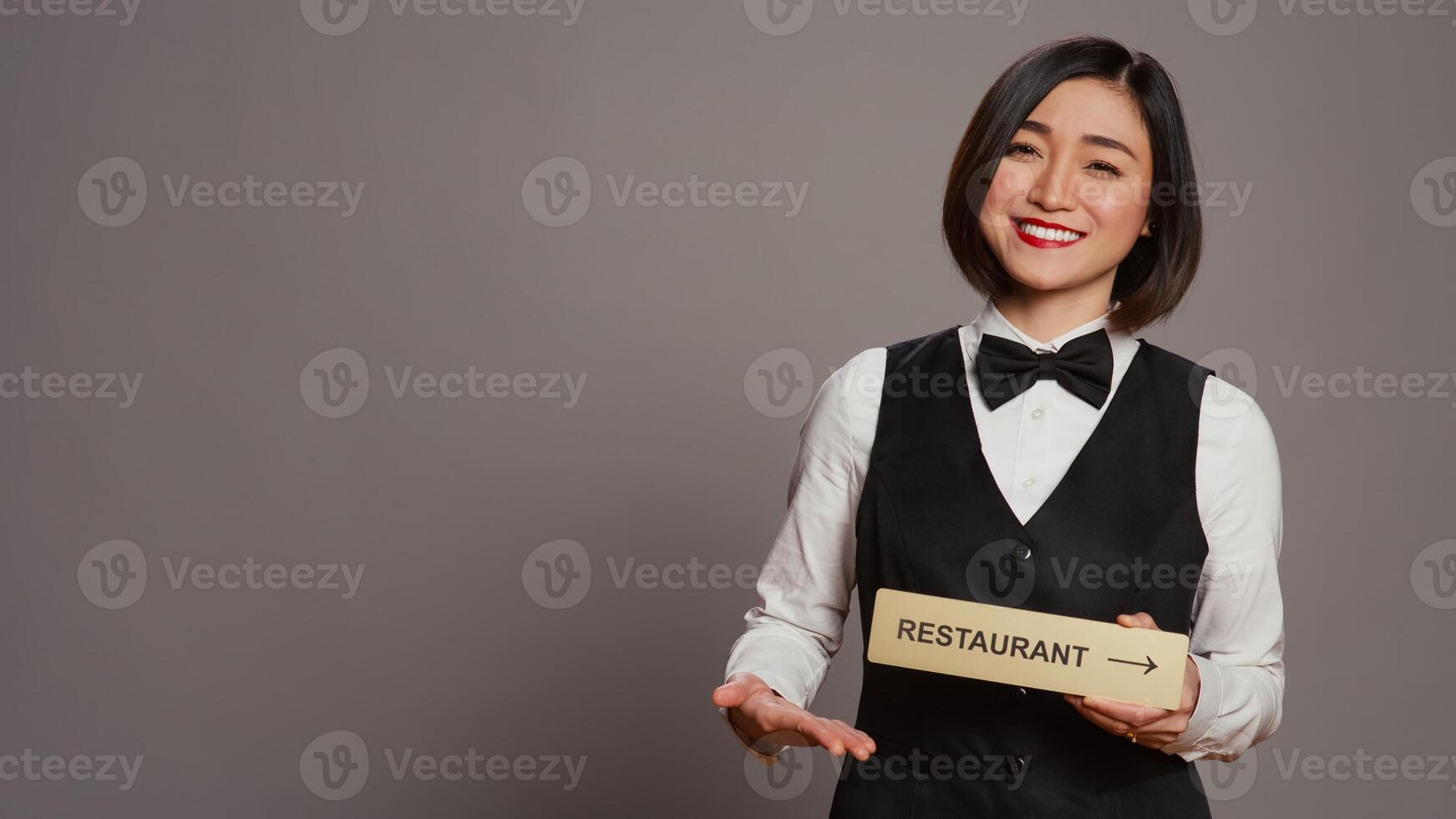 Asian hotel concierge holding restaurant sign to indicate direction, pointing towards dining area. Receptionist assisting clients to enjoy all amenities, stands over grey background. Camera B. photo