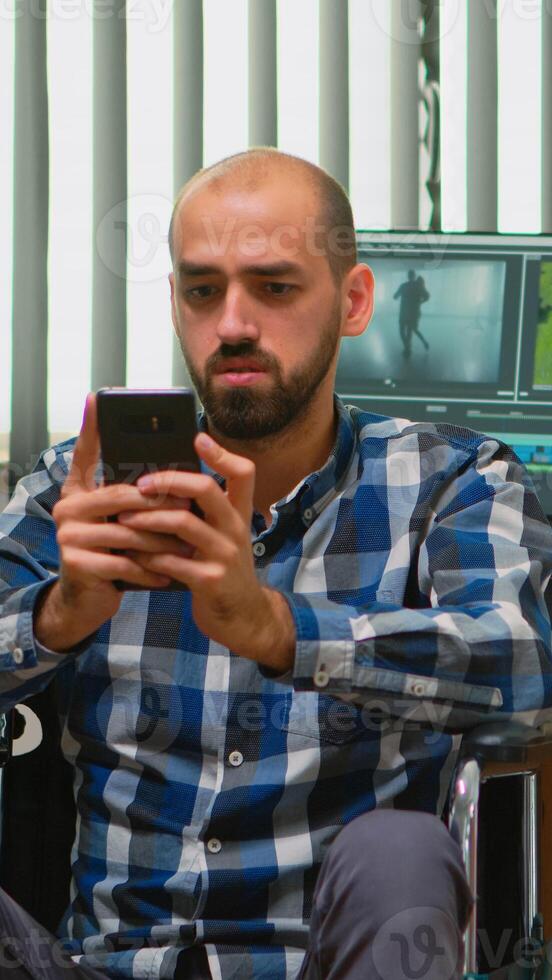 Freelancer photo designer with disability in wheelchair texting on smartphone taking break from editing video project creating content in modern company office. Blogger working from photo studio.