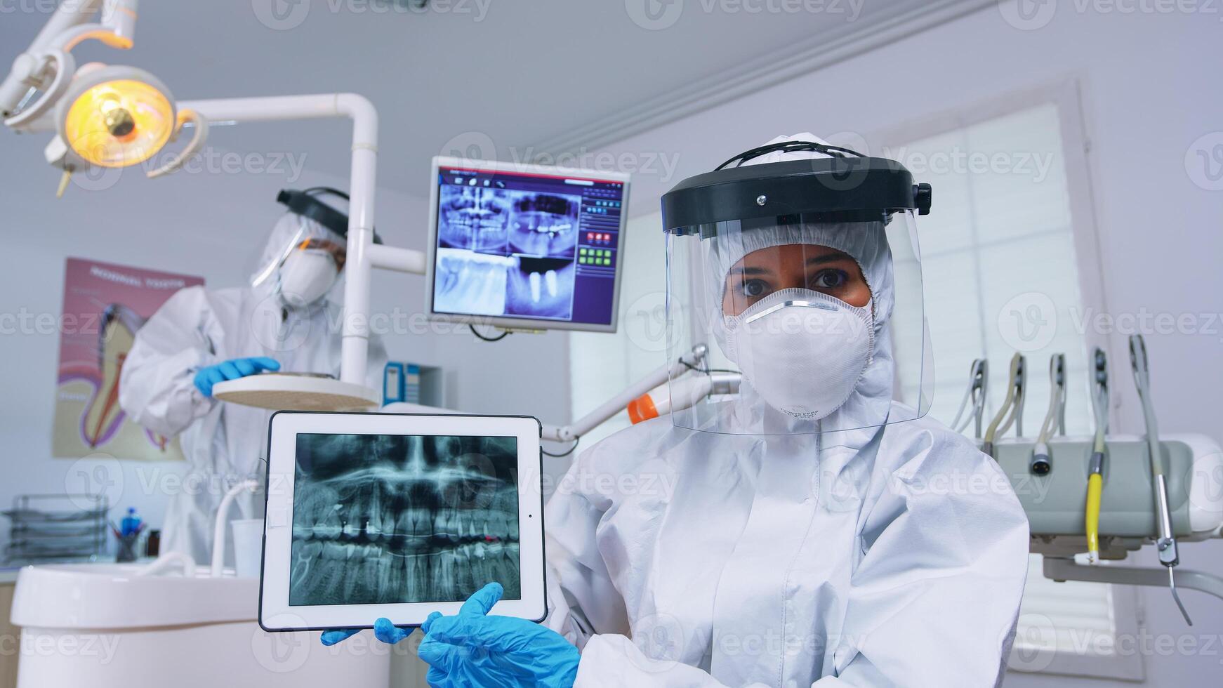 Patient pov in dental office with new normal discussing treatment of teeth cavity, dentist pointing on digital x-ray using tablet. Stomatology wearing protective hazmat suit against coroanvirus photo