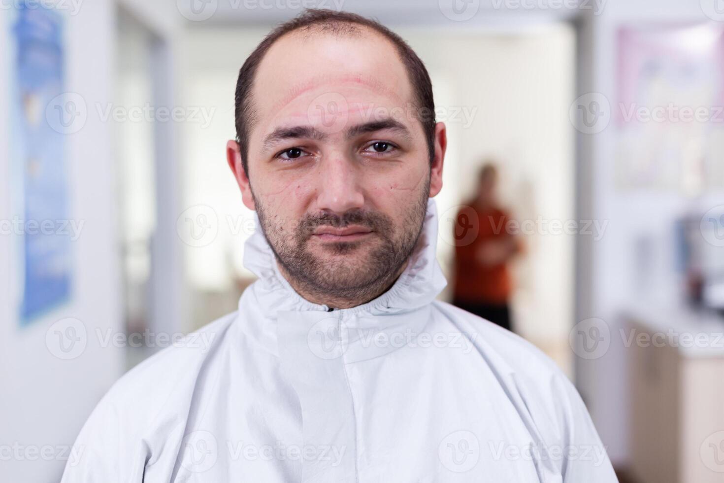 Portrait of exhausted doctor looking on camera wearing ppe suit without face shield sitting on chair in waiting room clinic. Concept of new normal in coronavirus outbreak. photo