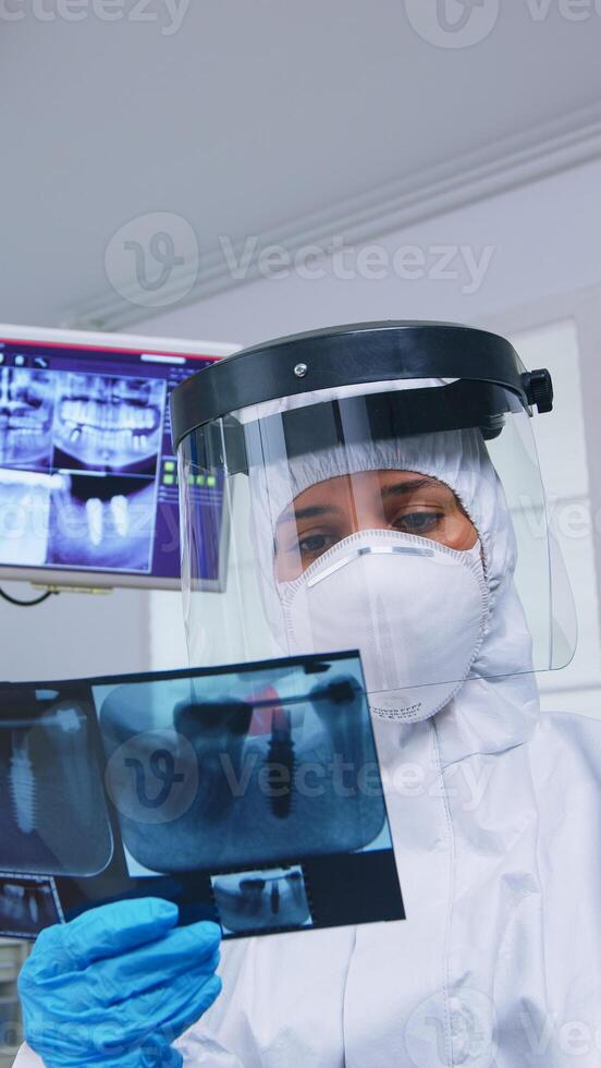Patient pov looking at dentist in ppe suit showing x-ray image in dental office. Stomatology specialist wearing protective hazmat suit against coroanvirus showing radiography in clinic with new normal photo