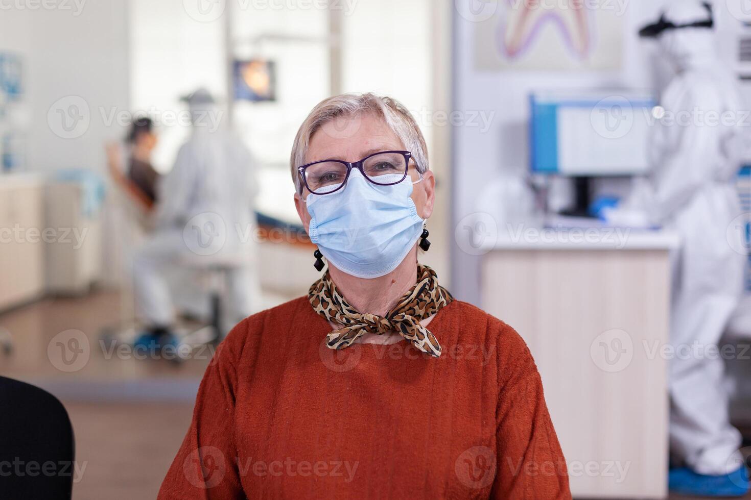 Portrait of retired patient in dental office looking on camera wearing face mask sitting on chair in waiting room clinic while doctor working. Concept of new normal dentist visit in coronavirus outbreak. photo