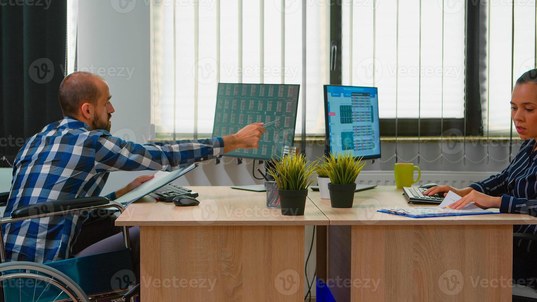 Handicapped paralysed businessman in wheelchair analysing economy data taking notes making financial expertise in business office discussing with colleague. Immobilized man using modern technology. photo