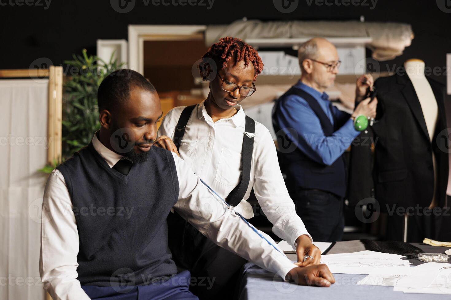 Suitmaker taking customer measurements for bespoken handmade business suit while elderly needleworker does adjustments on sartorial garment. Teamworking tailors manufacturing premium quality clothing photo