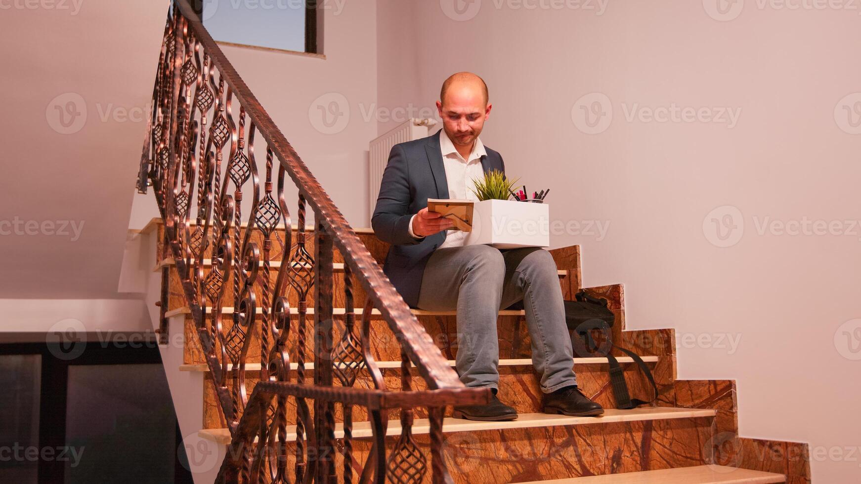Fired businessman sitting on stairs in corporate finance company holding box of personal employee stuff. Group of professional successful businesspeople working in modern financial building. photo