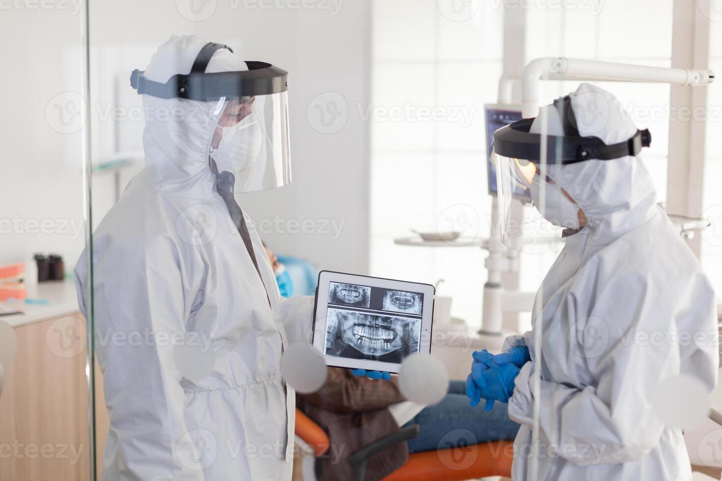Dentist doctors with ppe suit analysing teeth x-ray using tablet in dental room, planning surgery during global pandemic while patient waiting on stomatological chair. photo