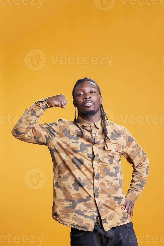 Portrait of athletic bodybuilder showing trained arm muscles in front of camera in studio over yellow background. African american man enjoying sport exercices, working out at triceps strength. photo