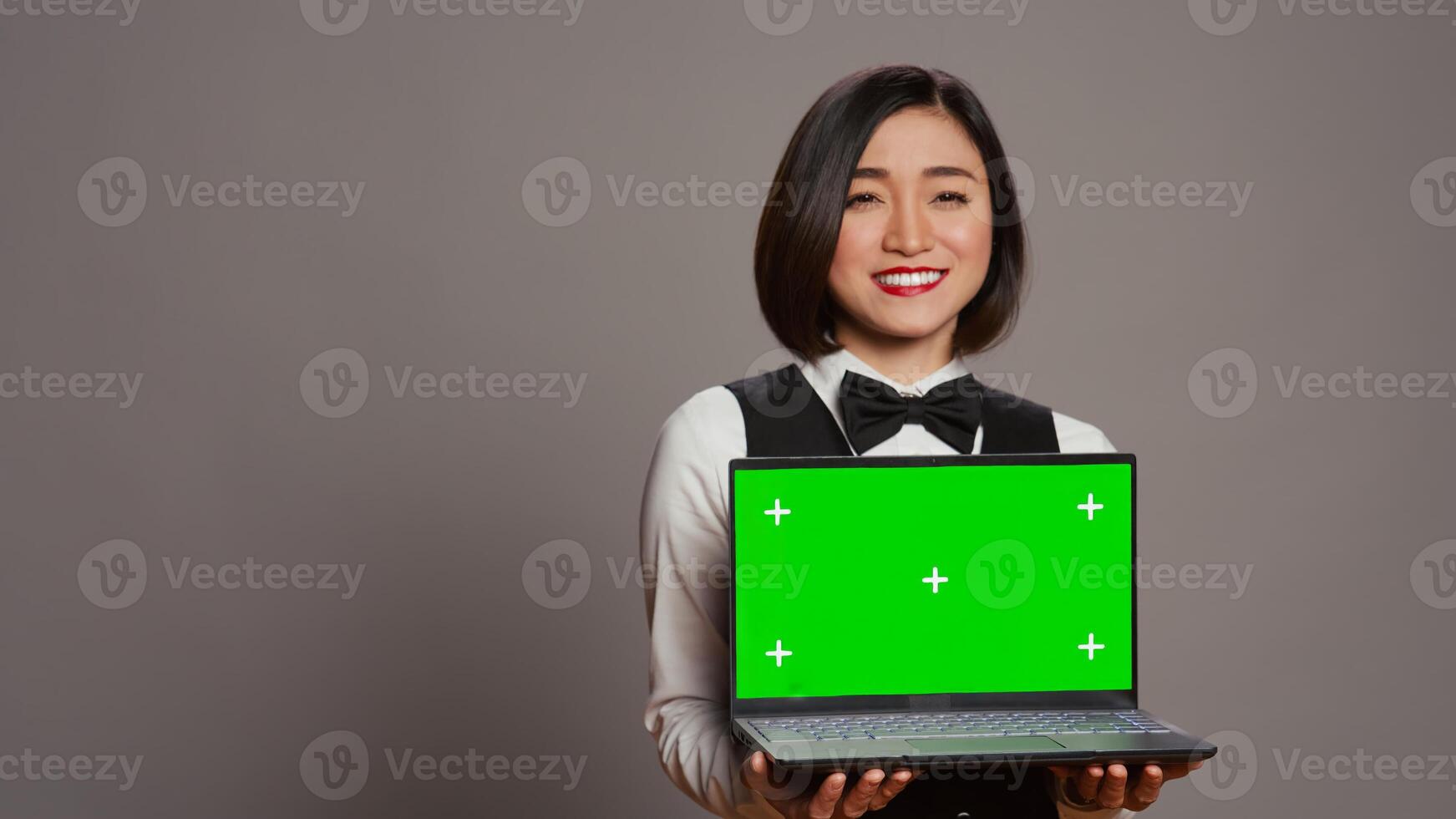 Asian hotel concierge holding pc with greenscreen on camera, presenting isolated mockup template on screen. Receptionist showing laptop with blank chromakey layout on display. Camera B. photo