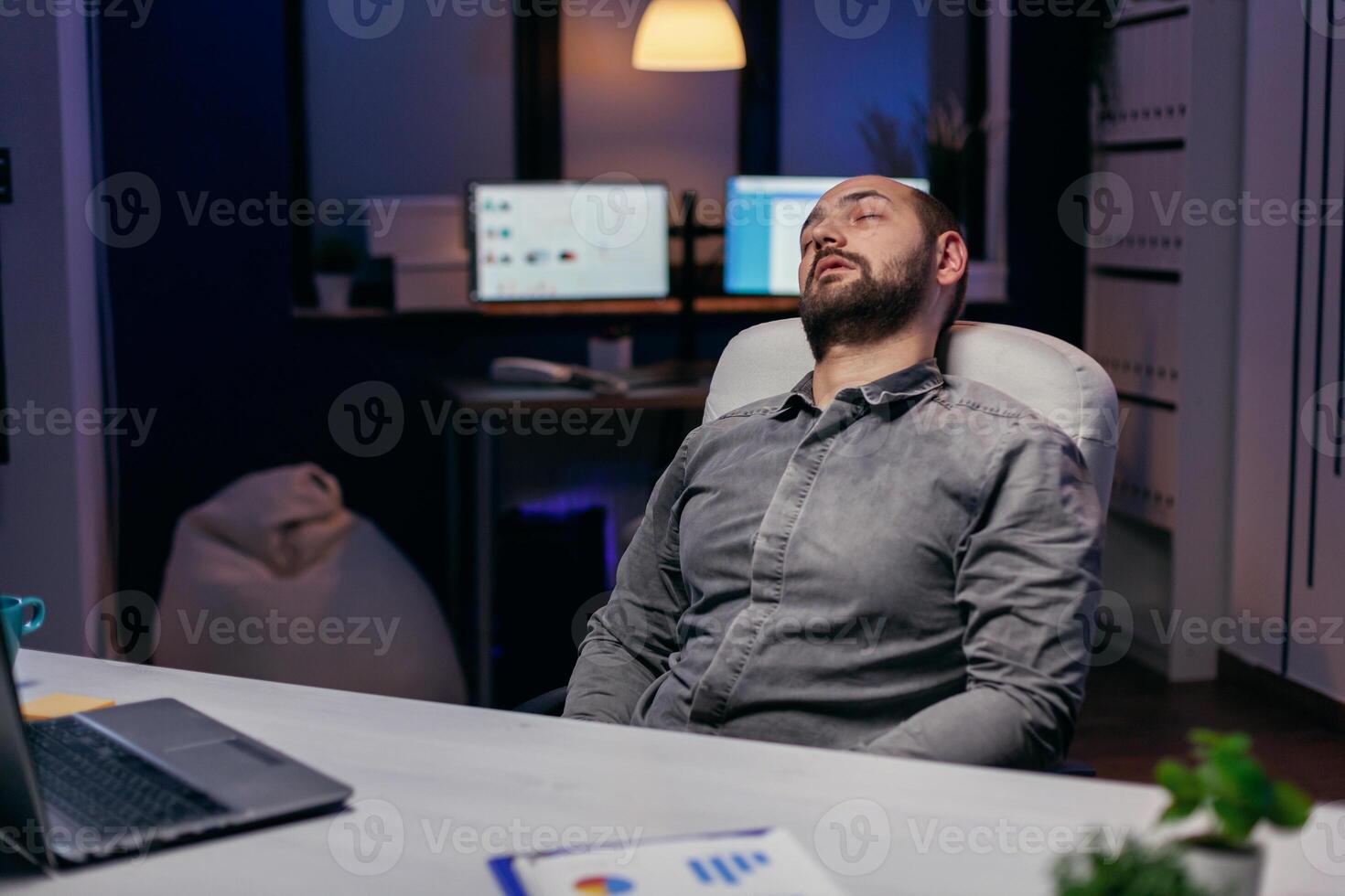 Young businessman resting on chair while working on deadline. Workaholic employee falling asleep because of while working late at night alone in the office for important company project. photo