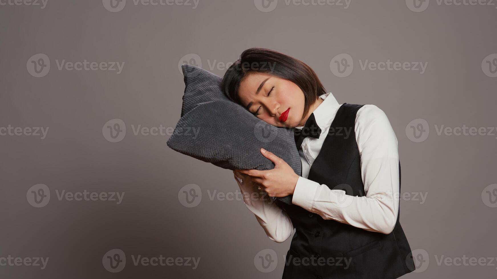 asiático frente escritorio personal que cae dormido con almohada en mano, sensación extremadamente cansado y con exceso de trabajo recepcionista siesta terminado gris fondo, agotado conserje con fatiga. cámara b. foto