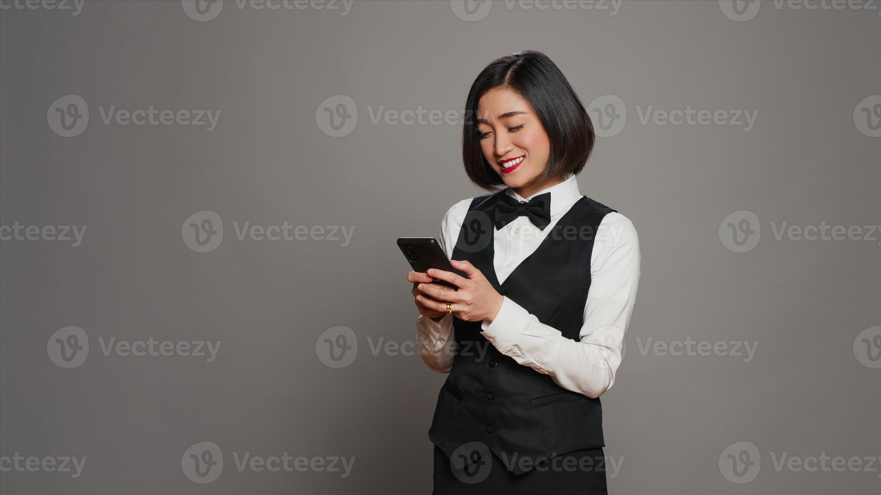 Hospitality industry worker using smartphone app over grey background, looking at online information to confirm room reservations. Asian receptionist browses websites. Camera A. photo