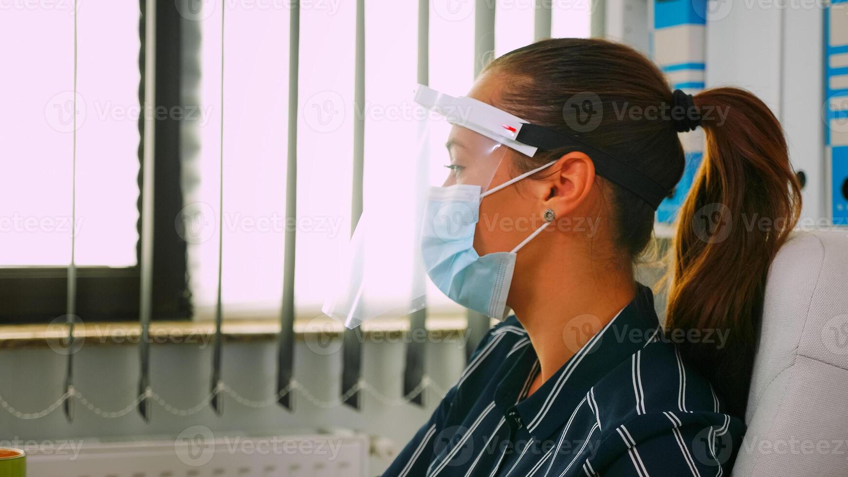 Portrait of businesswoman wearing protection mask and visor typing and talking with colleague working in new normal business office respecting social distance during global pandemic. photo