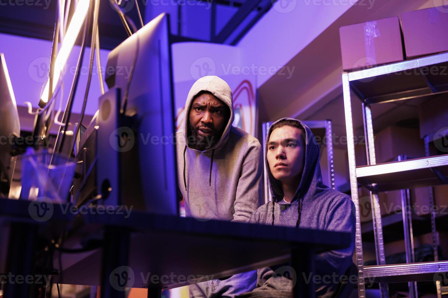 Two concentrated hackers collaborating to crack complex password and break into network system. Young diverse men in hoods working on computer together and coding virus malware photo