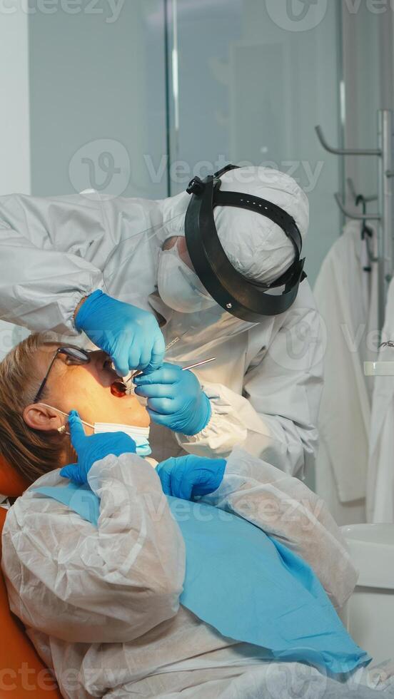Dentist with face shiled treating teeth to senior woman patient in dental clinic during covid-19 . Doctor and nurse working wearing coverall, protection suit, mask, gloves in stomatological office. photo