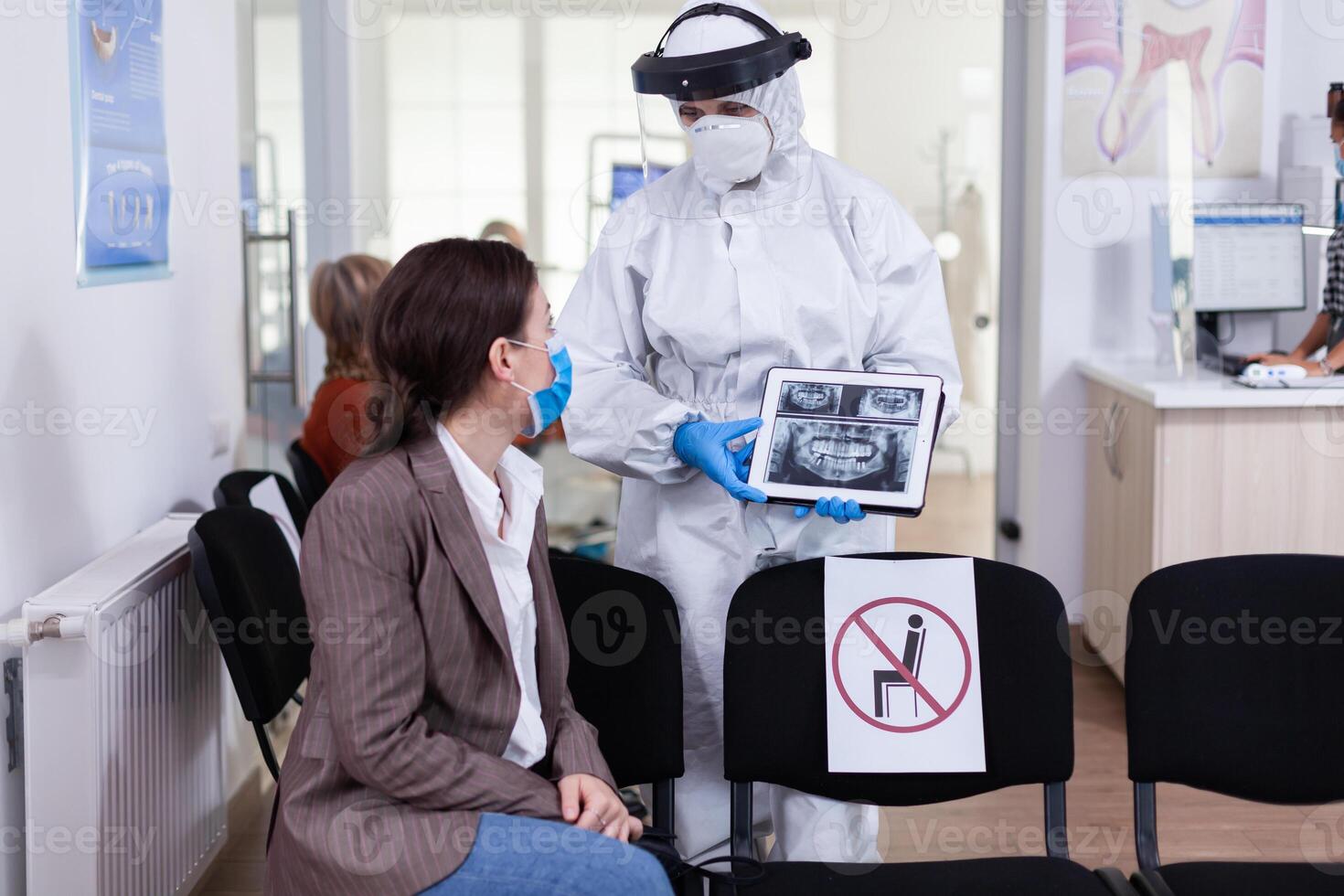 Stomatologist in protective suit pointing on digital x-ray of tooth explaining to patient treatment using tablet in covid-19 pandemic. Medical team wearing face shield, coverall, mask and gloves. photo