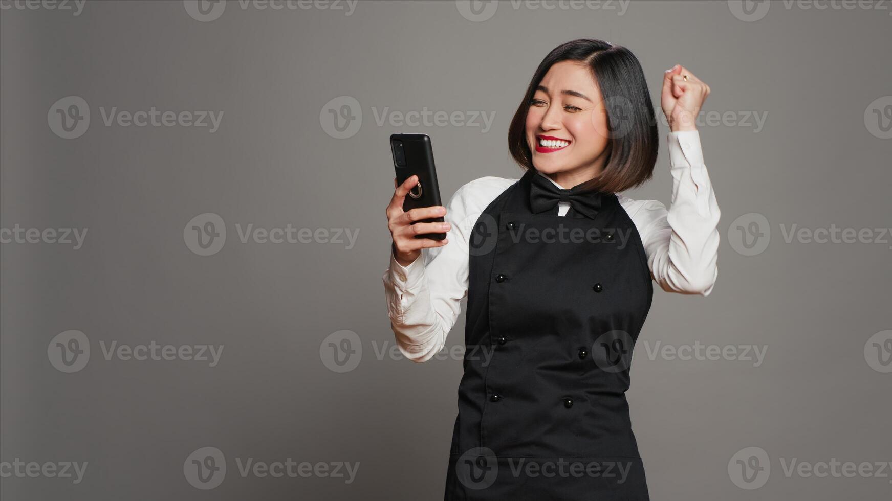 Asian server feeling happy after reading good news on phone, having a lot of clients with reservations for dinner. Waitress with apron using smartphone to see table bookings, enjoys work. Camera A. photo